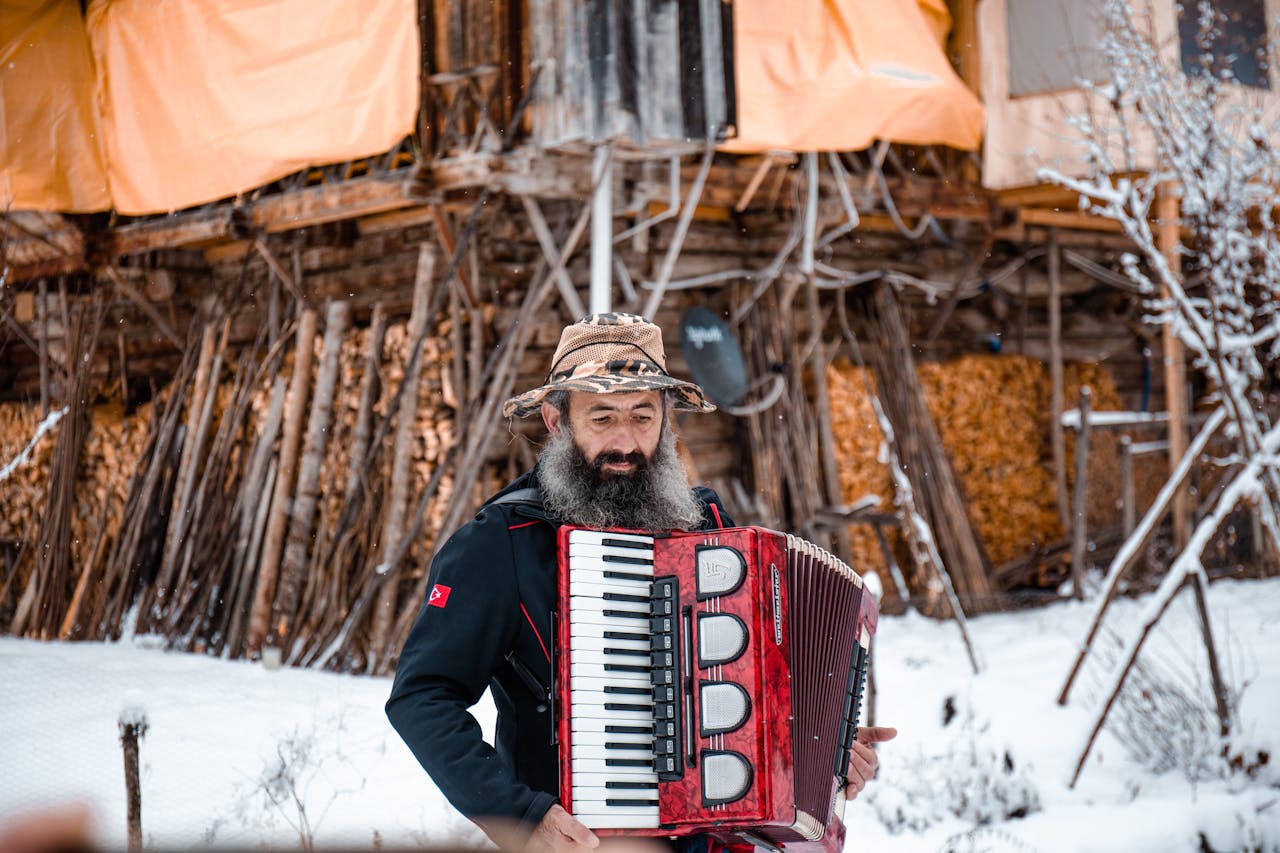 Bearded man plays accordion outdoors in snowy Şavşat, Turkey, with rustic background.