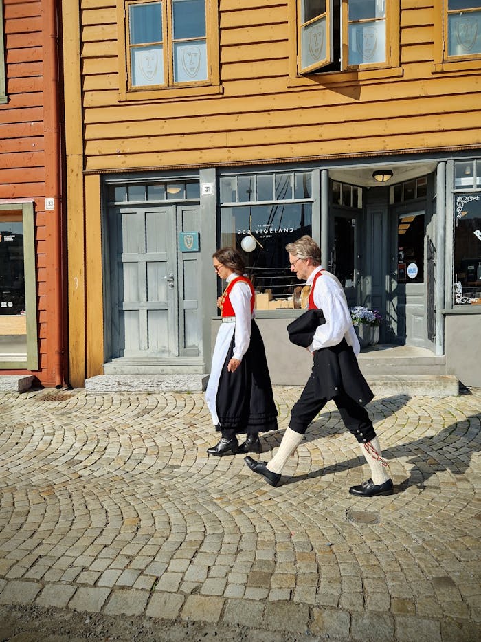 Couple in traditional attire walking down cobblestone street in Norway's picturesque town.