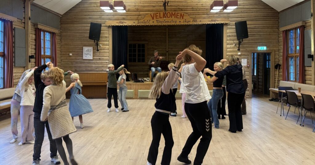 Born og unge dansar parvis i ein ungdomshussal medan ein spelemann speler hardingfele frå scena
